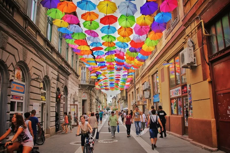 A vibrant pedestrian street in Timisoara, Romania, decorated with hundreds of colorful hanging umbrellas, with people walking and cycling past historic shops.