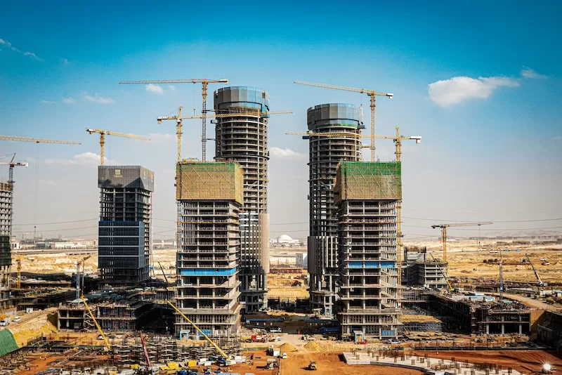 Multiple massive skyscrapers under construction in Egypt's new business districts, featuring several yellow cranes against a clear, bright blue sky.