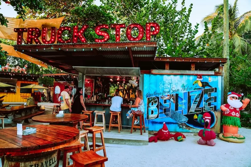A vibrant outdoor bar called Truck Stop in San Pedro, Belize, features a colorful mural, wooden tables, and festive decorations under a canopy of green trees. 