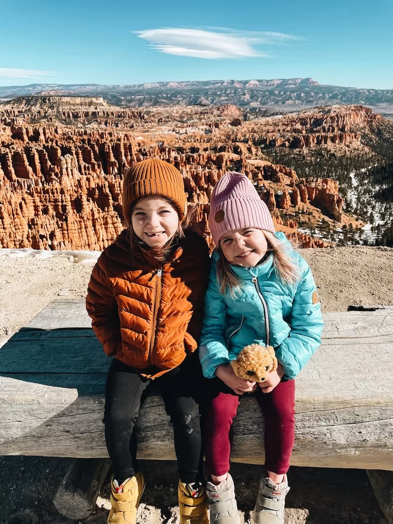 Two young girls, bundled in winter hats and jackets, sit on a wooden bench with a stuffed animal, smiling brightly against the stunning backdrop of Bryce Canyon National Park's unique hoodoos.