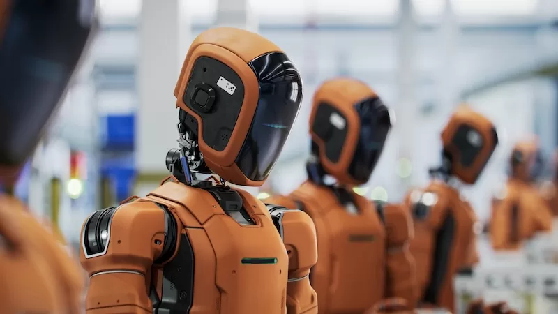 A line of advanced orange and black humanoid robots stands in a factory setting, with one in the foreground looking forward with a neutral expression. 