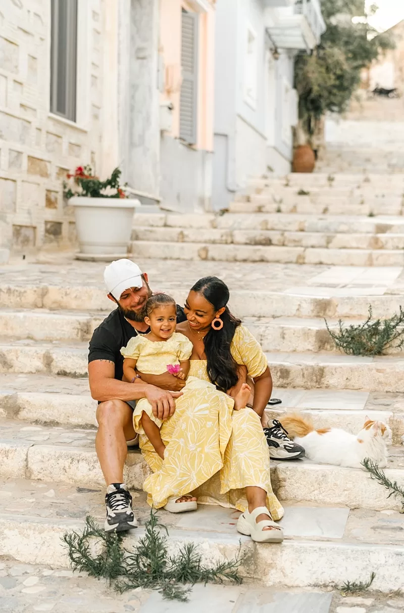A family sits together on stone steps in a charming European village, sharing a quiet moment of connection with a white cat resting nearby on the stairs.