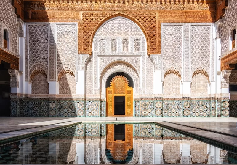 A serene Moroccan courtyard featuring a central reflecting pool, intricate zellige tilework, and beautifully carved plaster arches in Marrakech. 