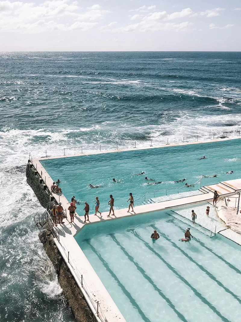 Aerial view of an outdoor swimming pool complex with turquoise water next to a rocky coastline and crashing ocean waves in summer daylight hours