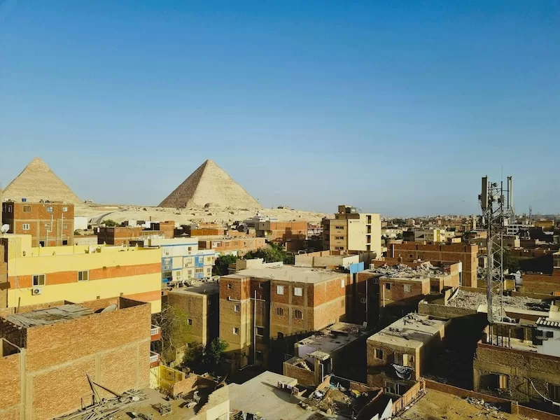An elevated view of a residential neighborhood in Giza with the iconic Great Pyramids rising majestically in the background under a hazy blue sky.