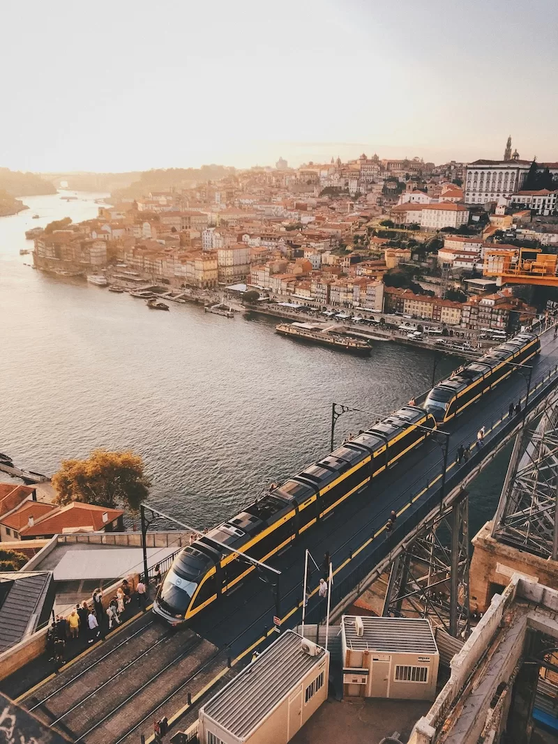 An elevated view of Porto, Portugal, at sunset, showing a train crossing the Dom Luís I Bridge over the Douro River, with historic buildings and orange rooftops lining the riverbanks