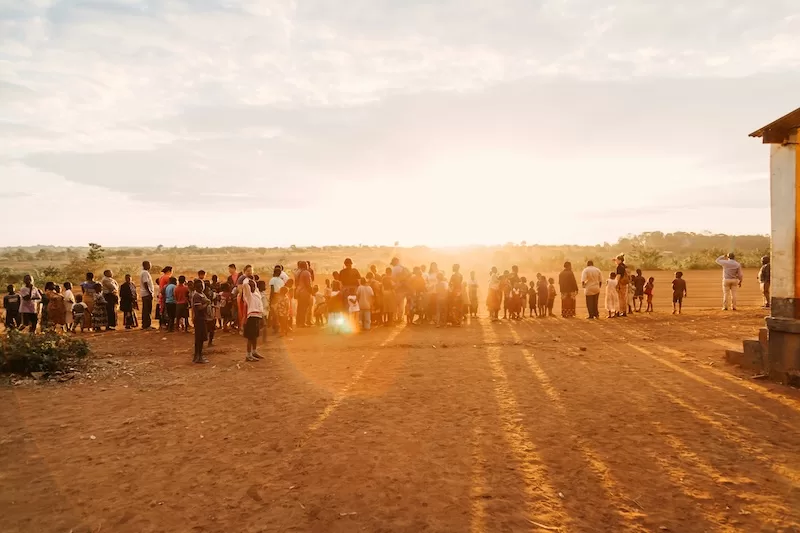 A long queue of people stands in a dry, dusty field under the bright, hazy sun in a rural African setting, waiting patiently outside a simple building.