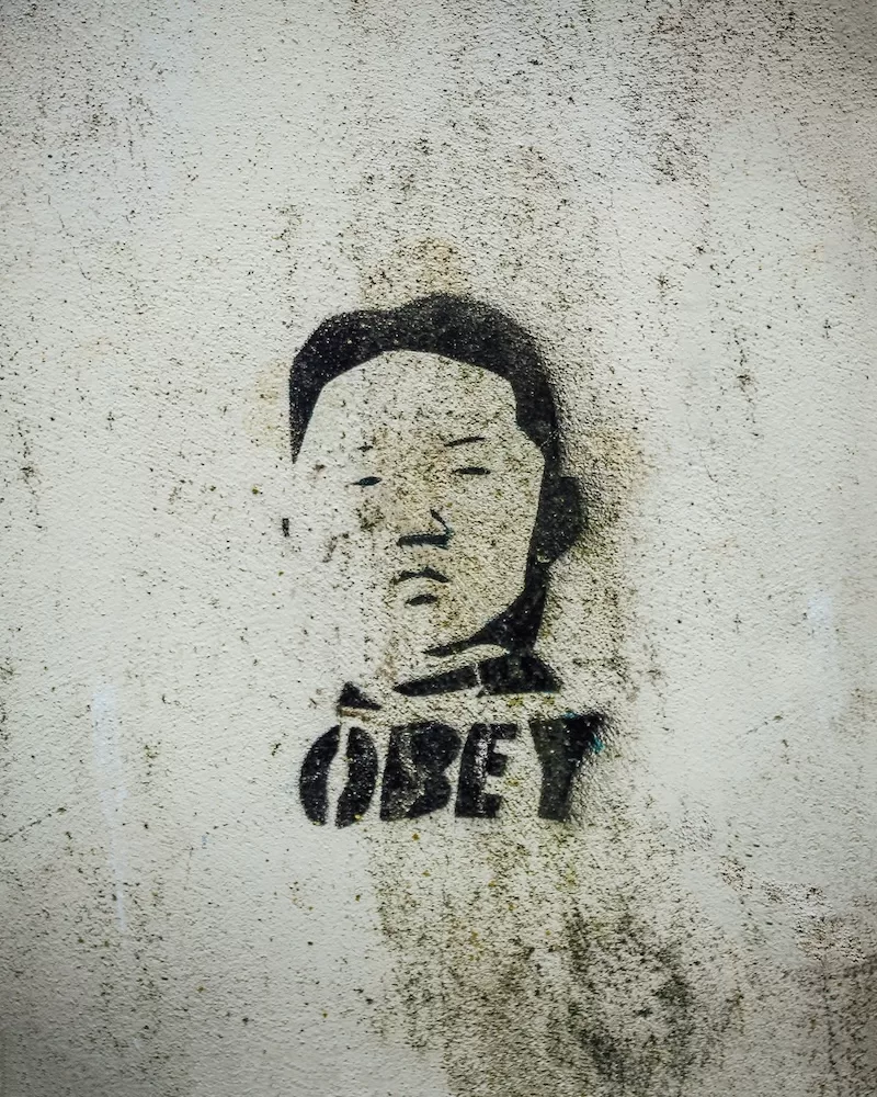 A faded black stencil graffiti of a political figure's face with the word "OBEY" underneath is seen on a weathered, dirty white concrete wall outdoors.
