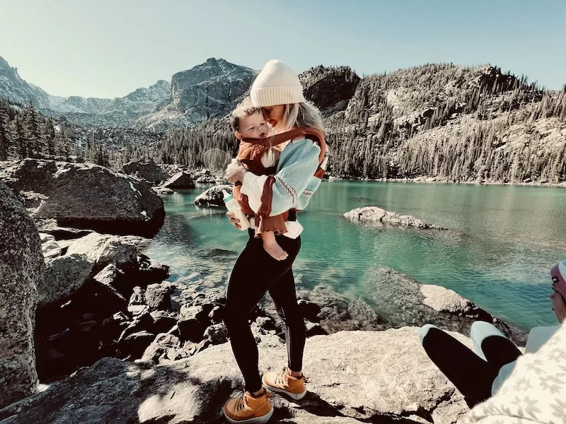 A mother carries her baby in a front carrier, walking along the rocky shore of a clear turquoise lake, with majestic snow-dusted mountains and a pine forest in the background. 