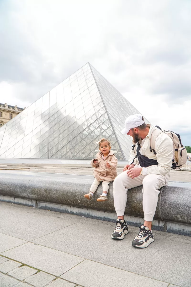 A father and his young daughter sit on a stone ledge in front of the glass Louvre Pyramid in Paris, enjoying a sunny day of exploration and travel.