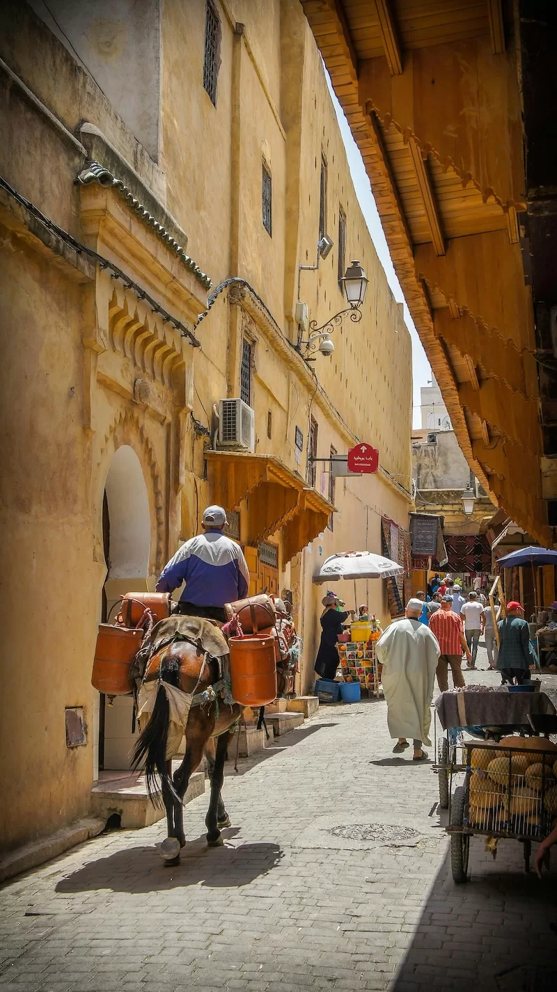 A man on horseback carries large orange containers through a narrow, sunlit alleyway in a bustling Moroccan medina filled with local residents. 