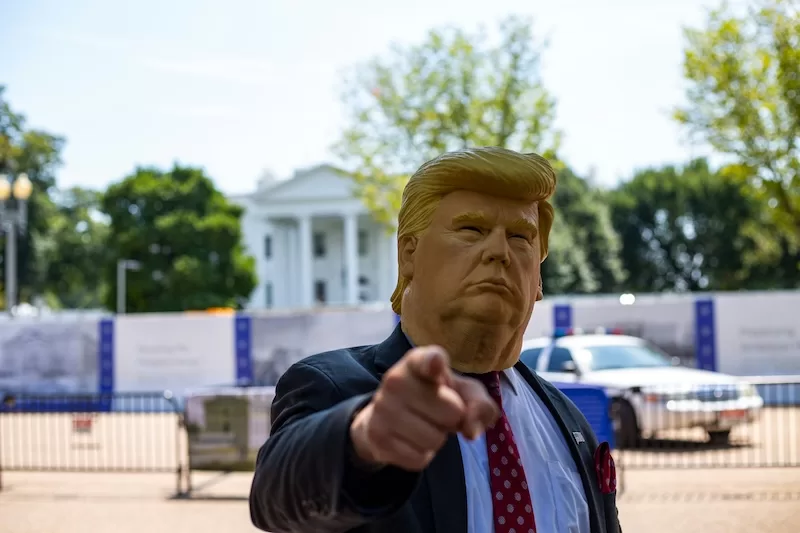 A person wearing a realistic Donald Trump mask and a suit points directly at the camera, with the White House blurred in the background on a sunny day.
