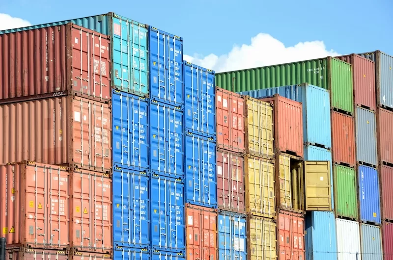 Stacks of colorful red, blue, yellow, and green shipping containers are piled high at a port under a bright blue sky with a few white fluffy clouds. 