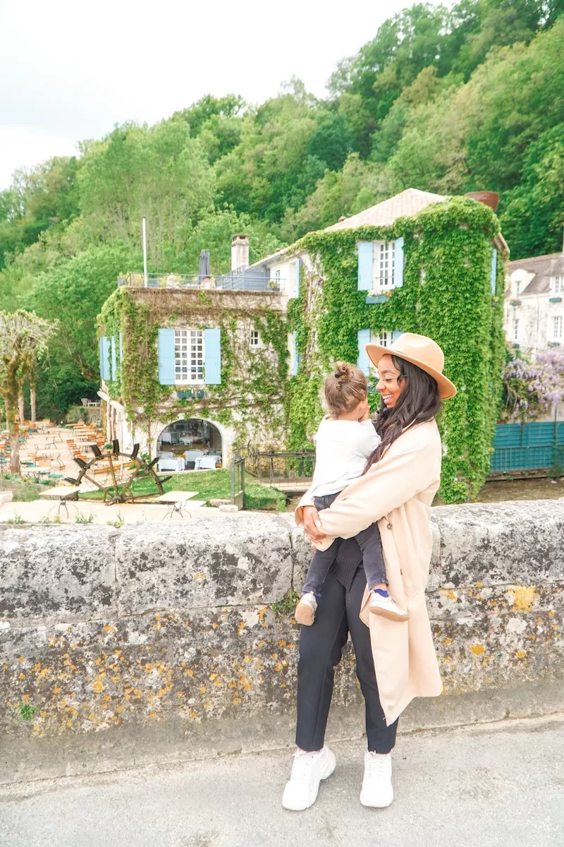 A mother holds her young daughter while standing on a bridge in a lush French village, with a historic ivy-covered building and waterwheel behind them.