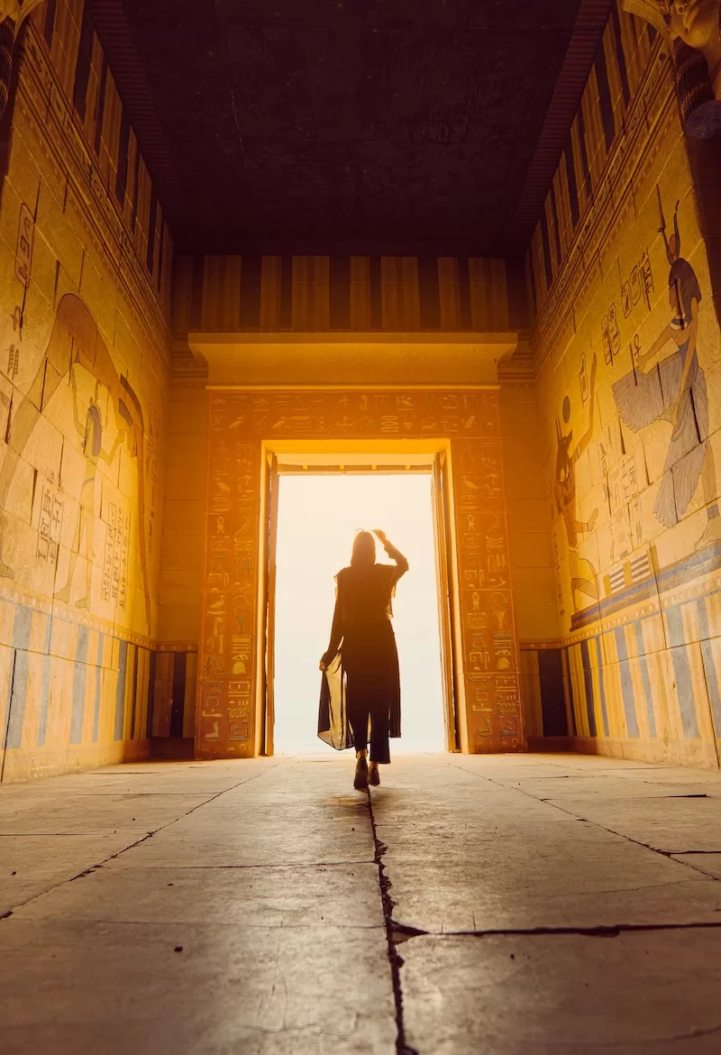 A silhouette of a person walking through a grand, sunlit ancient Egyptian temple doorway adorned with intricate hieroglyphics and stone carvings.