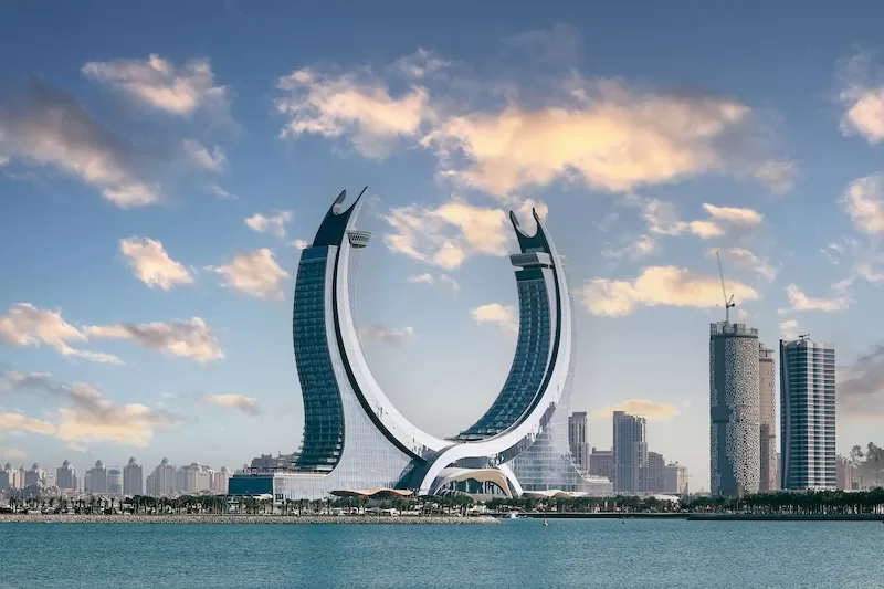 The iconic Katara Towers in Lusail, Qatar, stand majestically in a crescent shape against a blue sky with scattered clouds, overlooking the calm waters of the Arabian Gulf. 