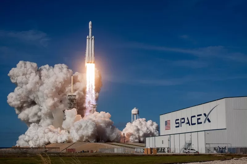 A massive SpaceX Falcon Heavy rocket lifts off from a launchpad, leaving a huge plume of white smoke and fire against a clear blue sky and a SpaceX building. 