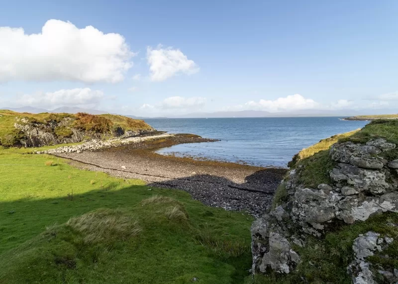 A tranquil view of a remote Scottish coastline with a pebble beach, green grassy hills, and calm blue water under a partly cloudy sky on a clear day