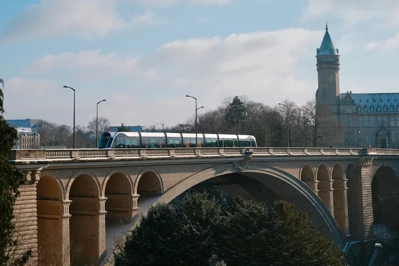 A modern blue and white tram crossing a historic stone bridge with arches over a river with a castle tower visible in the background landscape view