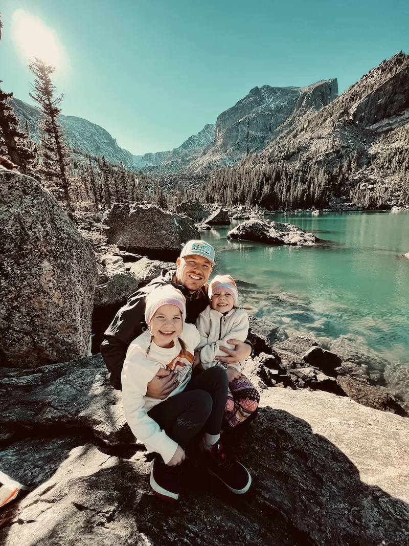 A father and his two young daughters pose happily on rocks beside a stunning turquoise alpine lake, surrounded by snow-capped mountains and dense pine forests. 