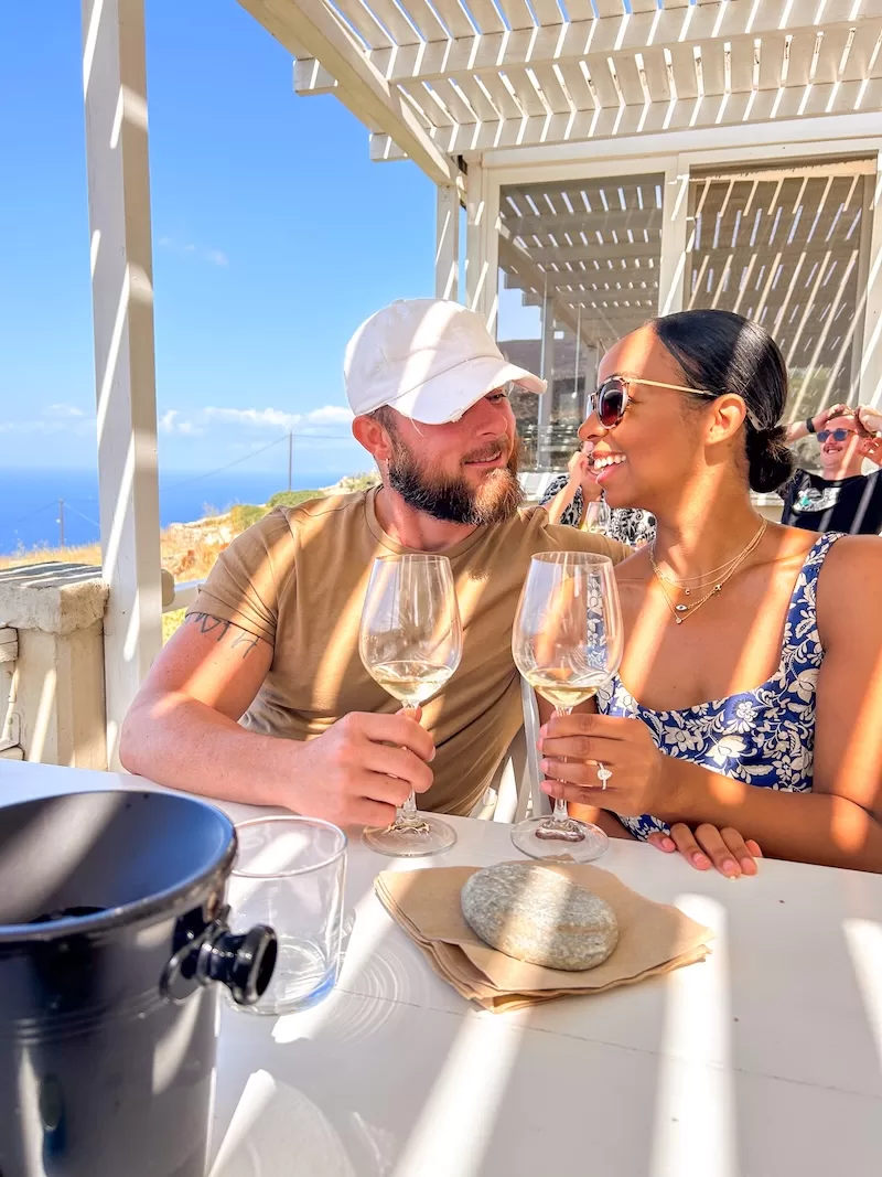 A couple shares a romantic moment at an outdoor restaurant in Greece, holding glasses of white wine under a white pergola with the blue sea in view.