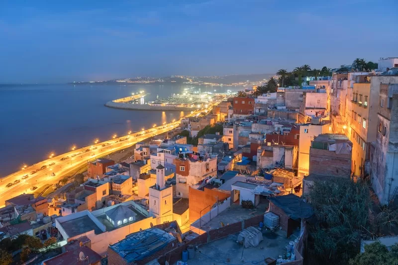 An aerial nighttime view of a coastal Moroccan city with lights reflecting on the water and a long, curved road stretching along the dark shoreline. 