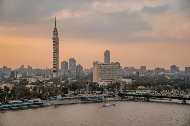 The Cairo skyline at sunset featuring the iconic Cairo Tower and modern buildings along the Nile River under a soft, hazy orange and grey sky.
