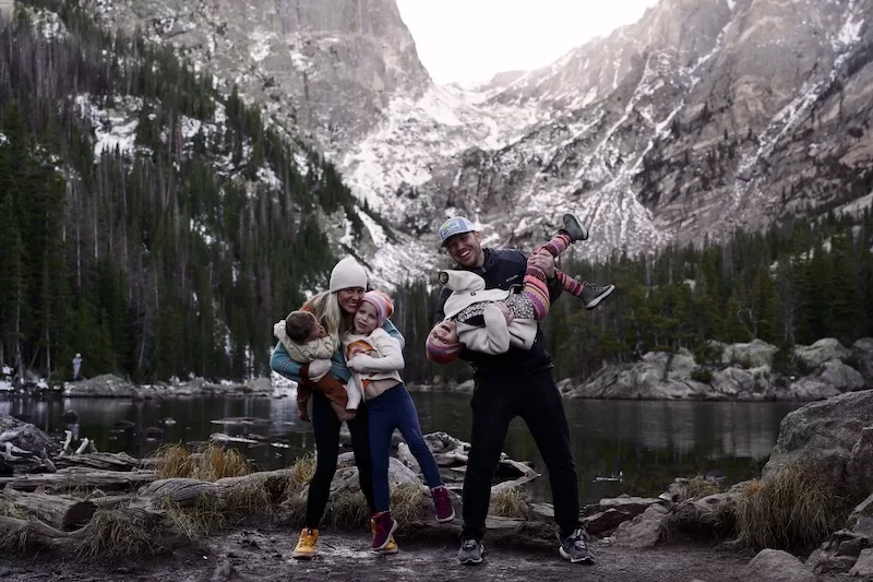 A joyful family of five, with parents playfully holding their children, stands on a rocky lakeshore, framed by a majestic snow-capped mountain range and a dense pine forest. 