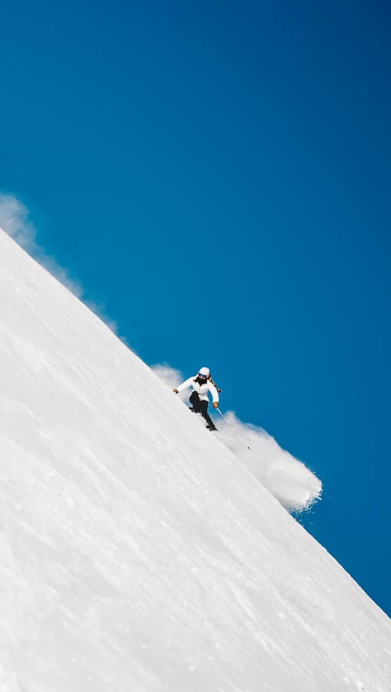 A skier in a white jacket carves down a steep, untouched snowy mountain slope at high speed, kicking up a cloud of fresh powder against a perfect blue sky. 