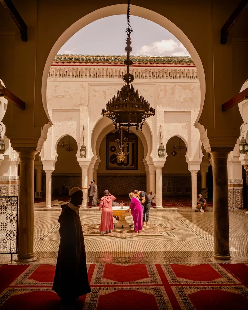 The interior of a historic Moroccan madrasa with people gathered around a central fountain beneath massive arches and an ornate chandelier.