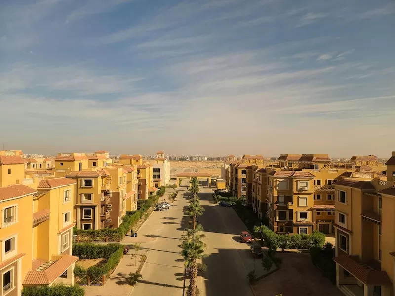 A modern residential complex in West Cairo with yellow buildings and red-tiled roofs, featuring a central palm-lined boulevard and a clear sky.