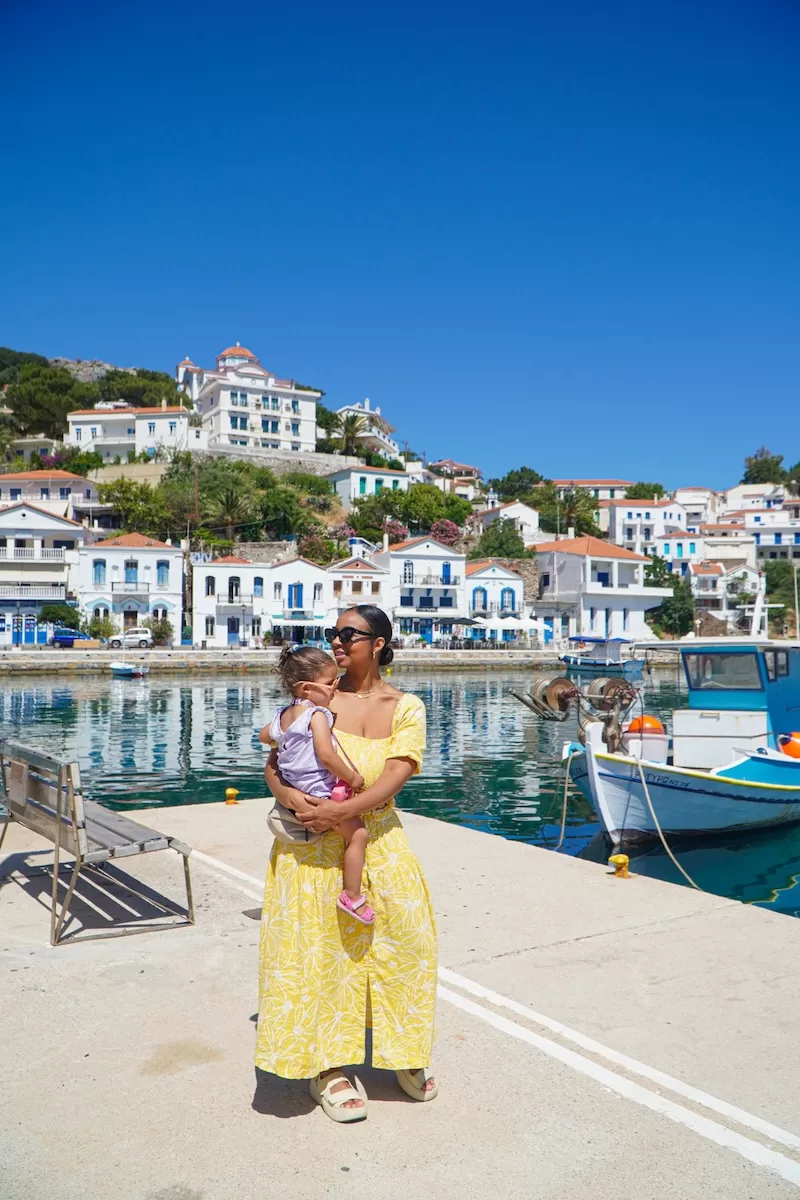 A mother carries her young child while standing on a pier in a Greek harbor, surrounded by traditional blue and white buildings and colorful fishing boats.