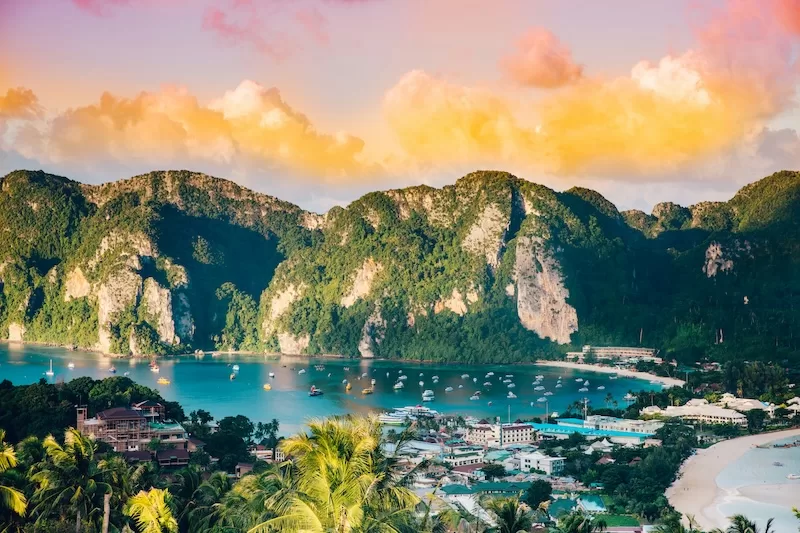 A breathtaking aerial view of Phi Phi Islands, Thailand, at sunset, showcasing dramatic limestone cliffs covered in lush greenery, a vibrant bay with boats, and a town nestled along the beach.