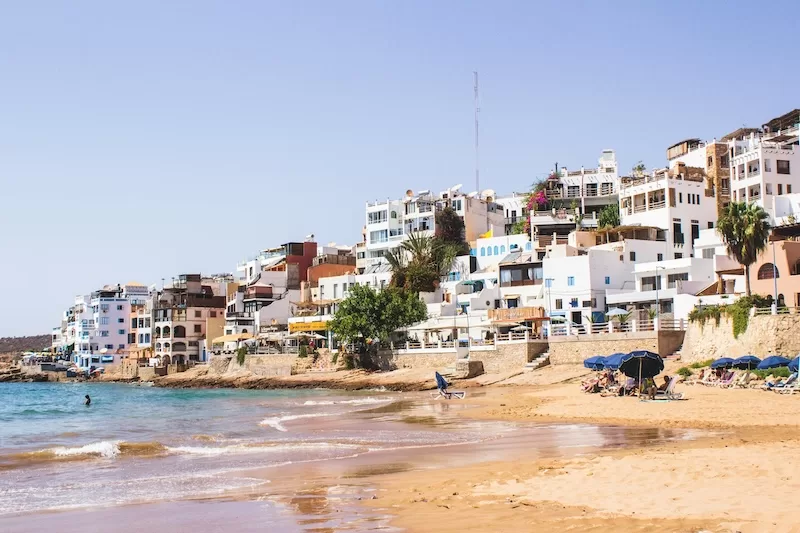 A row of white hillside buildings overlooking a wide sandy beach in Agadir, dotted with blue umbrellas and people enjoying the sunny Atlantic coast. 