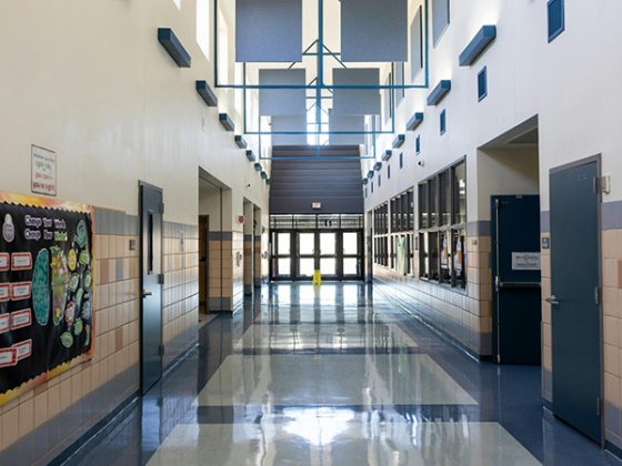 A modern school hallway with bright natural light from skylights, blue lockers, and glass doors leading to classrooms in a contemporary educational facility