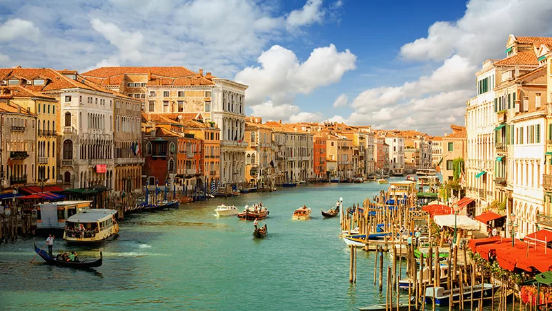 The Grand Canal in Venice with colorful historic buildings, gondolas, boats, and turquoise water under a blue sky with white clouds in the scenic Italian city