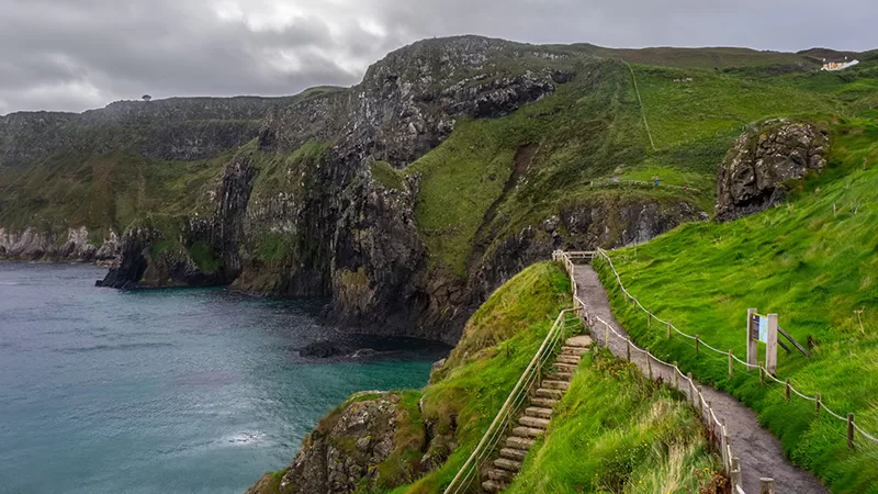 A dramatic coastal landscape with steep green cliffs, stone pathway, turquoise ocean water, and rocky outcrops overlooking a scenic seaside destination