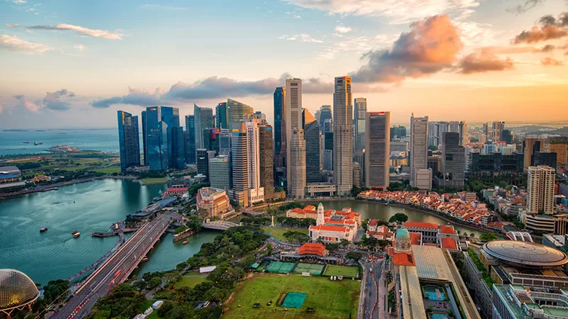 A vibrant cityscape at sunset showing a modern downtown skyline with tall skyscrapers, waterfront, and colorful clouds reflecting golden light over the city
