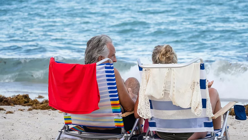A couple sits on a beach in front of ocean waves holding colorful towels, with the man wearing a hat and woman smiling while enjoying a sunny day