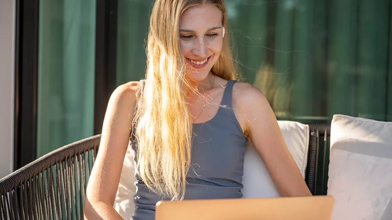 A blonde woman with long hair sits on a white outdoor lounge chair in front of green foliage, smiling while working on a laptop in natural sunlight