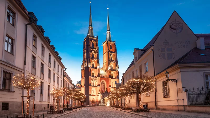 A historic European city street at sunset with ornate red brick cathedral towers, golden lighting, decorated trees, and traditional architecture buildings