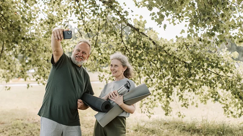 An older man and woman stand under green foliage trees holding a map, smiling and taking a selfie together during an outdoor adventure or travel day