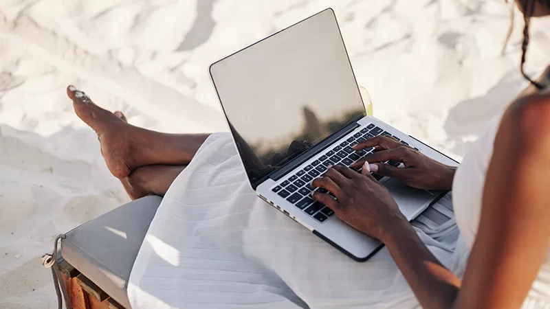A person working on a laptop while relaxing on a beach chair with bare feet visible, enjoying remote work in a tropical seaside vacation setting