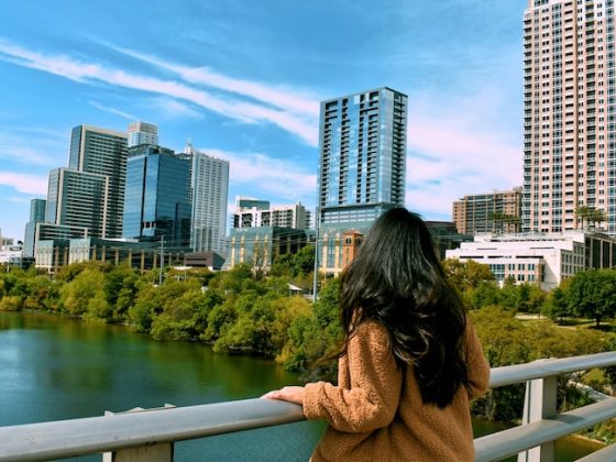 A woman with long dark hair looks out over a calm river toward a modern city skyline with tall glass skyscrapers under a bright blue sky with soft white clouds.