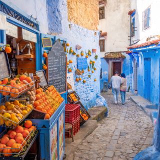 Chefchaouen, Morocco: Tourists passing by an orange juice shop in the narrow streets of Chefchaouen. Chefchaouen, located in northwest Morocco, is the capital of its namesake province. Renowned for its striking blue-colored buildings, it has earned the nickname "Blue City." Nestled in a mountainous region of northern Morocco, it lies between Tétouan and Ouazzane.