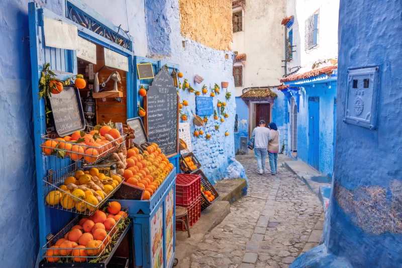Chefchaouen, Morocco: Tourists passing by an orange juice shop in the narrow streets of Chefchaouen. Chefchaouen, located in northwest Morocco, is the capital of its namesake province. Renowned for its striking blue-colored buildings, it has earned the nickname "Blue City." Nestled in a mountainous region of northern Morocco, it lies between Tétouan and Ouazzane.