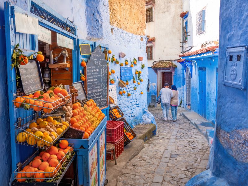 Chefchaouen, Morocco: Tourists passing by an orange juice shop in the narrow streets of Chefchaouen. Chefchaouen, located in northwest Morocco, is the capital of its namesake province. Renowned for its striking blue-colored buildings, it has earned the nickname "Blue City." Nestled in a mountainous region of northern Morocco, it lies between Tétouan and Ouazzane.