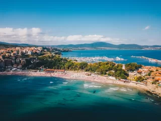 A woman with long dark hair looks out over a calm river toward a modern city skyline with tall glass skyscrapers under a bright blue sky with soft white clouds.