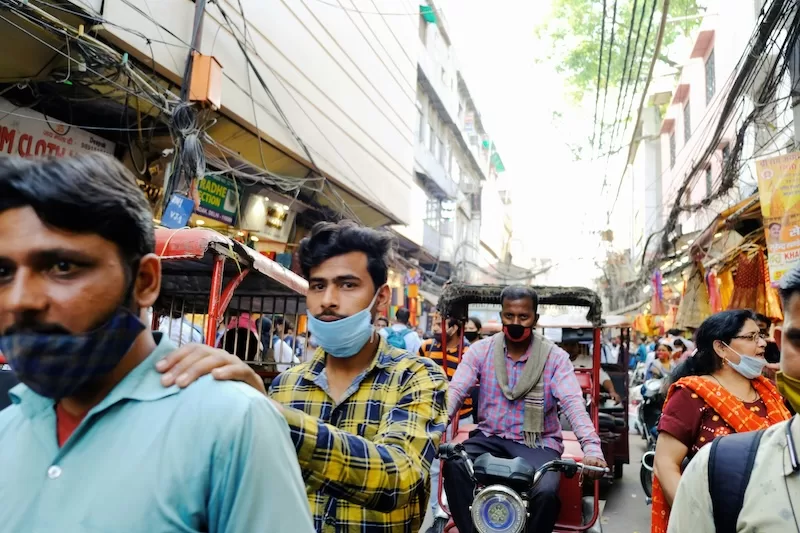 A crowded street in India with people wearing masks and riding rickshaws, illustrating the urban density and infrastructure challenges in cities like Delhi.