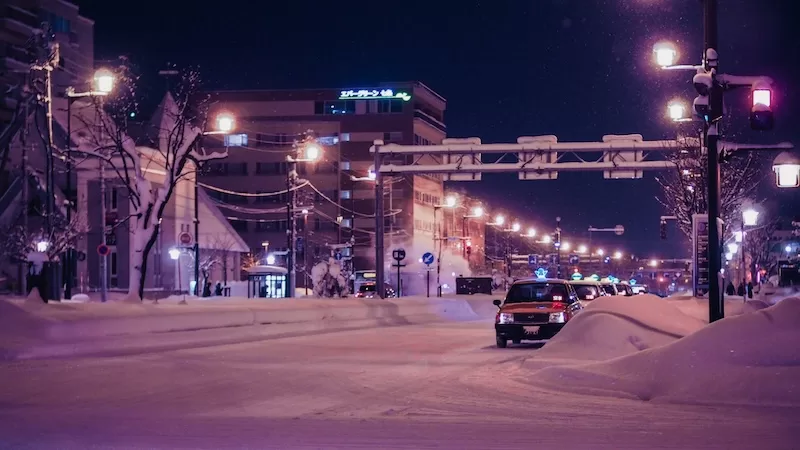 A row of taxis with glowing signs waits on a snow-covered street at night in a Japanese city, capturing the atmosphere of winter weather and urban transport.